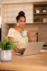 A woman at a kitchen table looking at a laptop with a warm cup of coffee